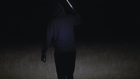 Rear view of unrecognizable man wearing hoody walking in darkness and illuminates his way with flashlight