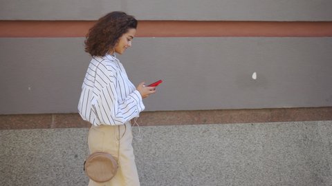 Smiling young latin woman with smartphone walking on urban city street against grey wall. Happy millennial Hispanic female with curly hair wearing earphones using mobile app listening music outdoor.