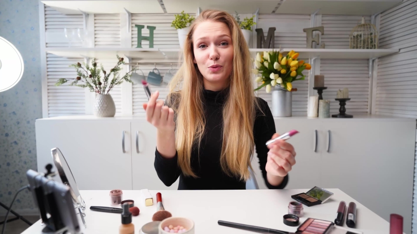 Vlogger female applies lipstick on lips. Beauty blogger woman filming daily makeup routine tutorial at camera on tripod. Influencer blonde lady live streaming cosmetics product comparison in studio. | Shutterstock HD Video #1057942774