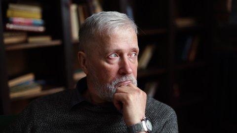 Close-up portrait of bearded gray-haired mature male, thoughtfully holding fist hand near chin, sitting at home on background of bookshelves in cozy dark room with an authentic aristocratic interior.
