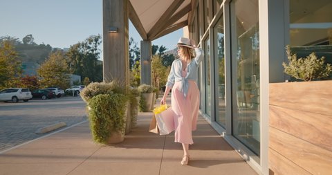 Attractive stylish woman in pink dress with paper bags walking by shopping mall. 4K slow motion girl shops at retail stores in the outdoor shopping village at sunny summer day. California, USA