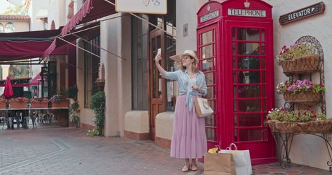 Cheerful smiling woman is having fun and making selfie on smartphone at red London telephone booth on a street of old town in European country. Stylish traveler is taking picture with London landmark
