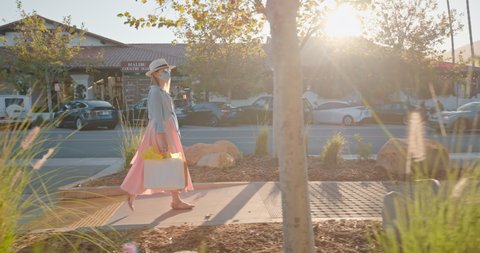 4K slow motion shopping during COVID-19. Young woman in face mask is walking at shopping village with sunset light on background. Stylish woman in pink dress with paper bags in shopping mall. USA