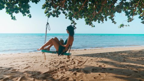 Carefree young woman on swing relax drinking coconut juice and enjoying sea on the tropical beach on summer vacation. Rest, travel concept