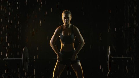 Portrait of Female performing deadlift exercise with weight bar. Confident young woman doing weight lifting workout barbell. Shot in a dark studio in the rain and staged light. Slow motion. Close up.