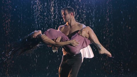 An attractive man holds and circles his beloved in the rain. Happy couple is enjoying each other. A love story in drops of rain. Shot in a dark studio with soft blue light. Close up. Slow motion.