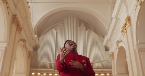 Afro american male performer in stylish red suit singing while standing at row of wooden pews in big hall. Talanted young guy performing gospel music emotionally and moving hands.