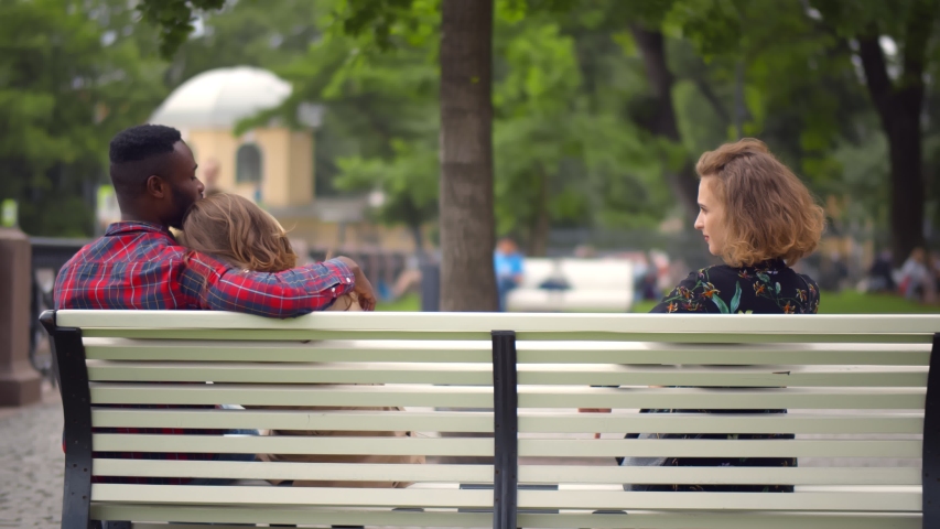 Cheating Boyfriend Hugging Girlfriend Holding Hands With Her GirlFriend Sitting On Bench Together In Park Outdoor. African guy secretly holding hand of other woman