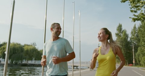 Happy couple running near water, tracking shot of delighted sportsman and sportswoman running on embankment near water during fitness training on summer day, slow motion