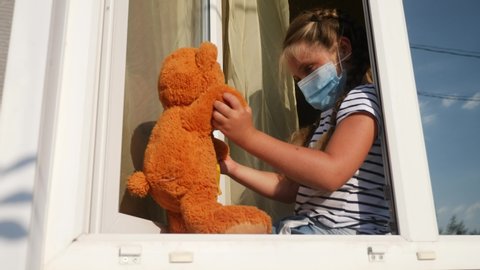 bored kid in medical mask in home quarantine coronavirus sitting by the window covid 19. child with a toy teddy bear in protective mask looking out the window. coronavirus epidemic prevention concept