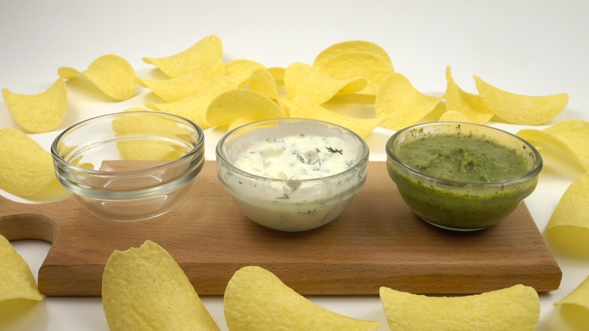 Thick red sauce is poured into a glass cup. Various sauces stand among potato chips