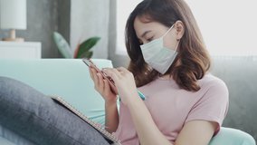Young Asian woman wearing protective face mask while working at home in living room. New normal healthy lifestyle at home concept. 