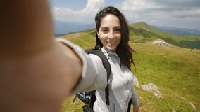 A carefree female tourist with backpack is making a selfie or technology video call to friends or relatives just reached a peak while hiking in the middle of hills surrounded by green nature.