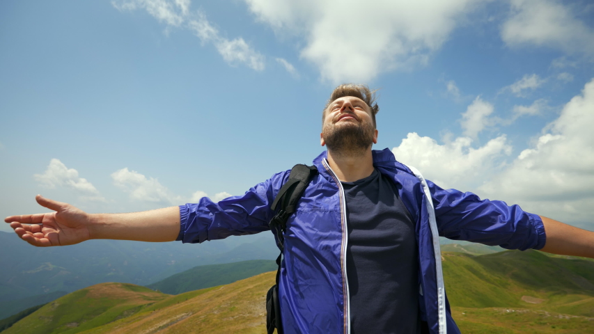 A carefree happy male tourist with backpack is breathing deeply with open arms raised just reached a peak while hiking in the middle of hills surrounded by green nature. | Shutterstock HD Video #1057973167