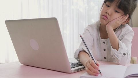 Asian homeschooling girl kid feeling bore to do homework and assigned job from remote school teacher. Child making boring face when working on computer on table. Remote online education concept.
