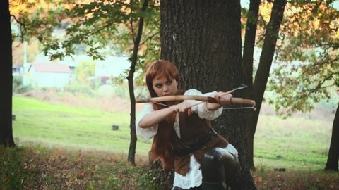 A woman archer hides near a tree and aims an arrow from a bow. Medieval hunter in a dense green forest. Сostume of a vintage warrior with a weapon in his hands. Fantasy shooting, video footage