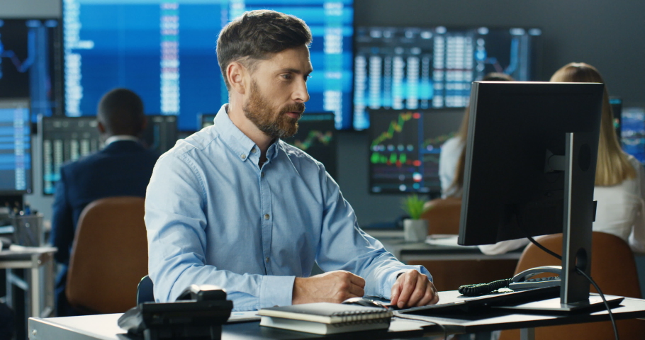 Portrait of young Caucasian handsome man trader working at computer, looking at camera and smiling in trading office of stock market. Happy cheerful male successful broker with smile. Businessman. Royalty-Free Stock Footage #1057979554