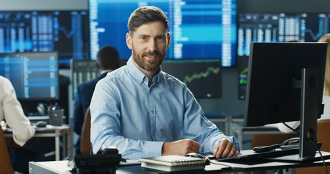Portrait of young Caucasian handsome man trader working at computer, looking at camera and smiling in trading office of stock market. Happy cheerful male successful broker with smile. Businessman.