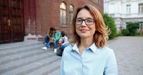 Close up portrait of beautiful Caucasian female teacher in glasses standing in front of school and smiling to camera. Mixed-races junior students sitting and using tablet on background. Study concept