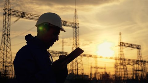 An electrical engineer wearing a helmet works with a tablet near high voltage electrical lines at sunset. A working electrician works with the documents of an electrical substation. business supply of