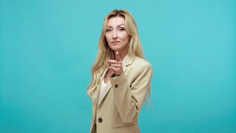 Angry middle aged blond woman in business suit touching nose and pointing finger to camera, showing liar gesture, suspecting and blaming in lying. Indoor studio shot isolated on blue background