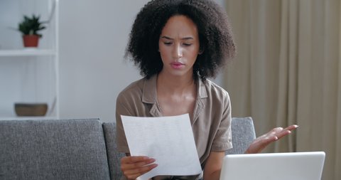 Young foreign African woman sitting on couch checking mail, receiving eviction letter, debt notice from tenant landlord, feeling annoyed desperate fear, worried, hopes for solution, problem concept