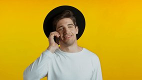 Young man in white wear and hat talking on mobile phone on yellow background. Trendy hipster guy have conversation. Smartphone, technology concept.