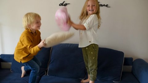Pillow fight. Mischievous preschooler children jumping on a sofa and hitting each other with pillows. Boy and girl play together. Active games for siblings at home.