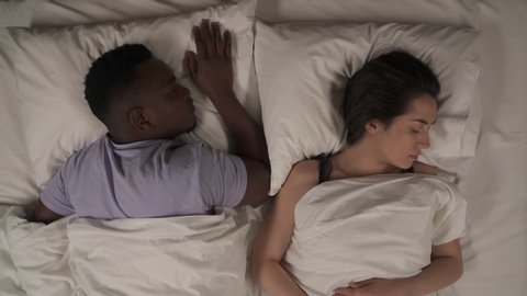 Close up of mixed-race couple in bed at night. African man sleeping quietly on his stomach. Caucasian woman tossing and turning in dream. Sleep disturbance