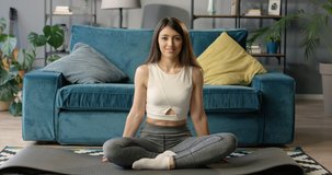 Portrait of smiled beautiful Caucasian young brunette woman sitting in lotus pose on yoga mat and doing namaste gesture in cozy livin room.  