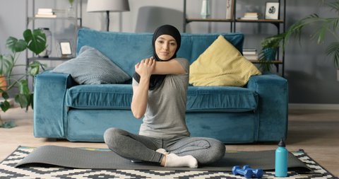 Muslim young beautiful woman in black traditional hijab sitting on floor on yoga mat and warming up arms. Exercising at home.  