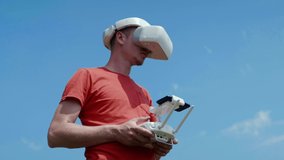 Man controls drone through remote control and looks at the video with goggles on head. Drone operator in red T-shirt in park shoots video. Blue sky on the background