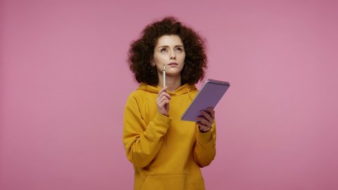Thoughtful creative young woman afro hairstyle in hoodie thinking over future business plan, taking notes in paper notebook, writing creative idea and smiling inspired happy. indoor isolated