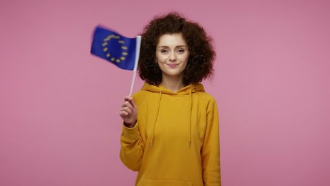 Happy student young woman afro hairstyle in hoodie waving European Union flag and smiling, cheering democratic laws, human rights and freedoms in Europe. indoor studio shot isolated on pink background