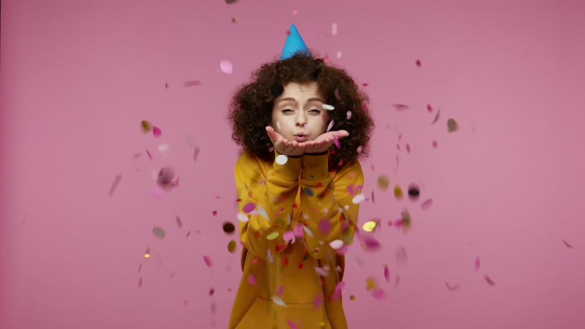 young woman afro hairstyle in hoodie with funny cone on her head laughing, moving in dance and blowing confetti glitters, celebrating birthday congratulating on anniversary. studio shot isolated Royalty-Free Stock Footage #1058012833