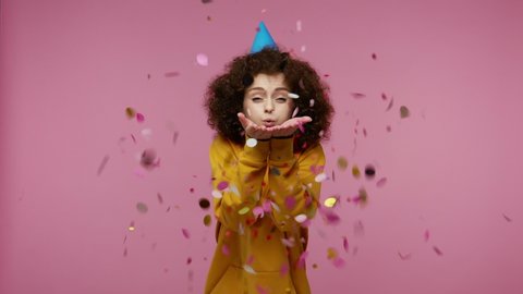 young woman afro hairstyle in hoodie with funny cone on her head laughing, moving in dance and blowing confetti glitters, celebrating birthday congratulating on anniversary. studio shot isolated