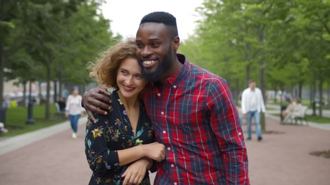 Beautiful diverse young couple in love walking in summer park. African man hugging smiling caucasian girlfriend walking together in city park enjoying romantic date