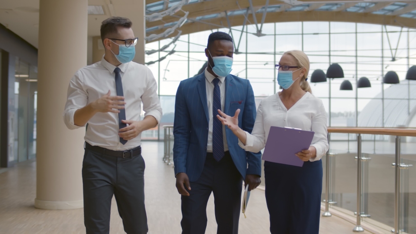 Business team wearing protective masks walking in modern office corridor. Diverse colleagues in safety mask during epidemic discussing project walking in hallway Royalty-Free Stock Footage #1058014219