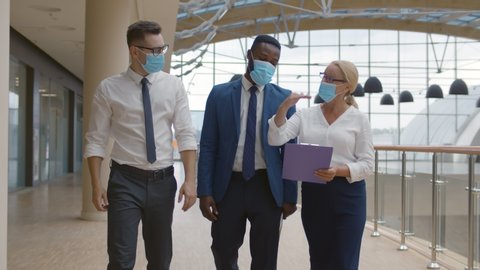 Business team wearing protective masks walking in modern office corridor. Diverse colleagues in safety mask during epidemic discussing project walking in hallway
