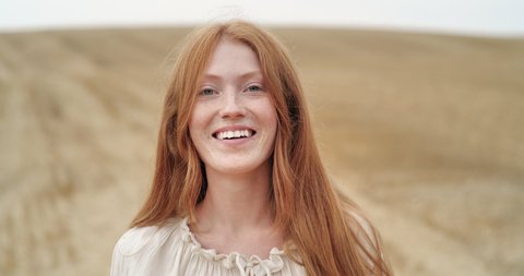 Cute Ginger Woman with long Red Hair walking on the Field, looking at Camera. Happy Ginger Girl is spinning ti the Camera with happy Smile, Summer Face Female. Emotions. People. Portrait.