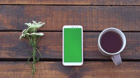 Closeup top view flatlay 4k video of cute wild flowers, mobile smartphone with blank green screen and cup of green tea isolated on brown wooden background. Woman touches phone with one finger.