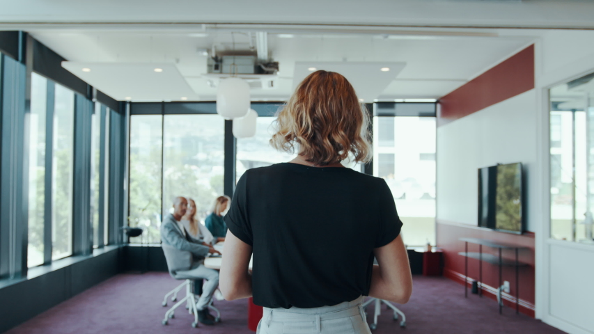 Rear view of a female entrepreneur walking to the conference table with colleagues.  Entrepreneur arriving in the conference room with her team settling down around the table.
 | Shutterstock HD Video #1058030677