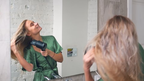 Charming smiling lady with light hair in green silk dressing gown looking at her reflection in bathroom mirror and drying her hair with hair dryer in morning
