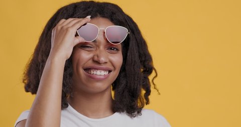 Playful black girl raising sunglasses up and winking, flirting on camera, orange studio background