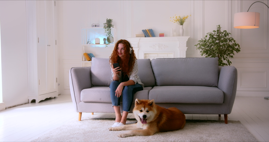 Funny young woman with red curly hair sitting on couch listening to music through headphones and petting her Akita Inu Dog. Pet is licking woman