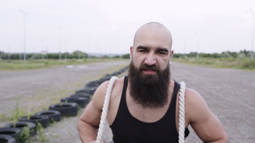 Athletic Guy Goes In For Sports. Man Runs And Pulls Car Tires Tied To A Rope. He Frowns At The Heavy Load.