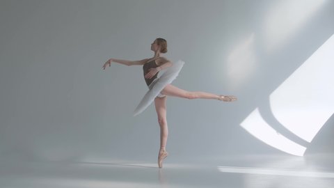 Professional ballerina in a white tutu dances in a large training hall. Girl does dance steps in the stage costume. Shot on a white background in the spacious and brightly lit studio. Slow motion.