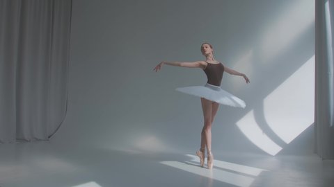 Female dancer does ballet exercises in stage dress with open back. Rehearses dance moves in the spacious studio bathed in sunlight. Slow motion.