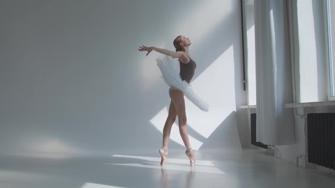 Young ballerina in stage dress with open back rehearses training in the studio standing by the window. Flexible girl on a white background in bright light. Slow motion.