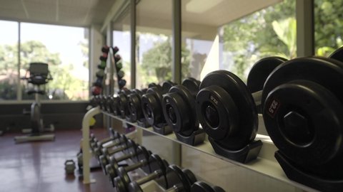 Handheld, forwarding shot of barbells sitting on rack inside a large gym. Selective focus.